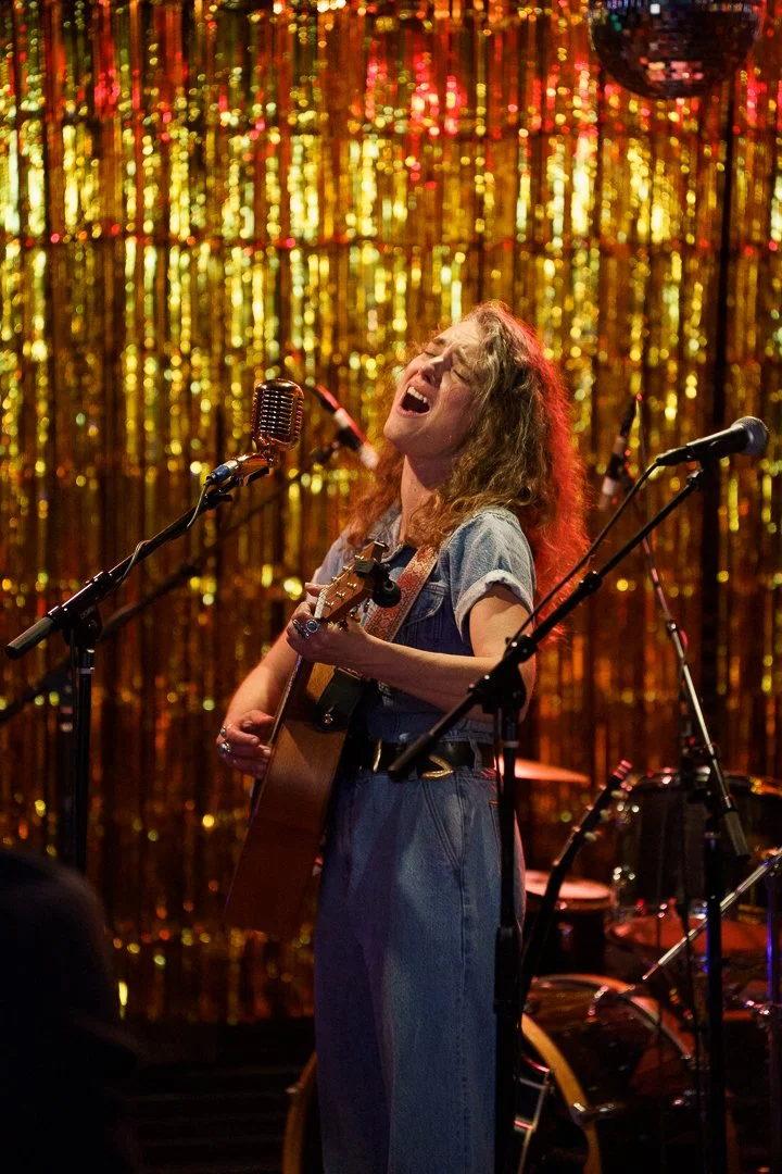A woman singing passionately while playing an acoustic guitar on stage with a golden sequin backdrop.