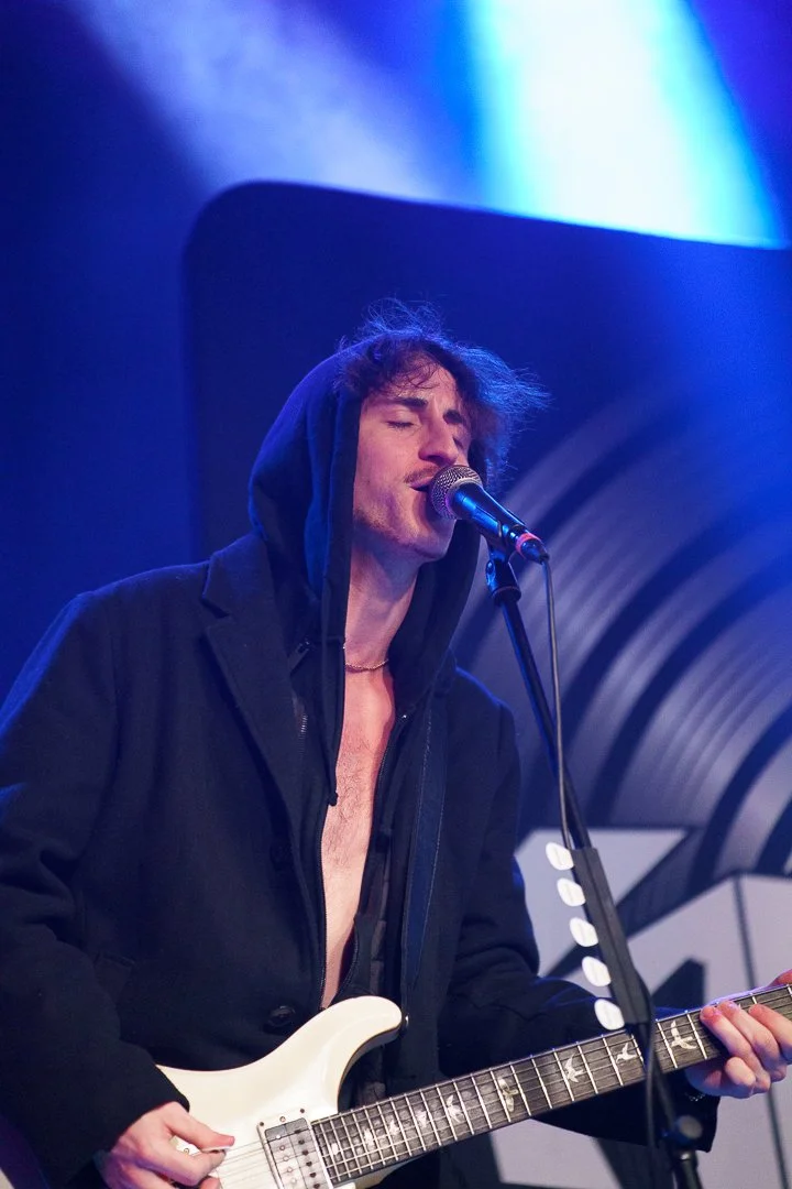 A male musician with dark, tousled hair, dressed in a black hoodie with the zipper half down, playing an electric guitar and singing into a microphone on stage with blue lighting.