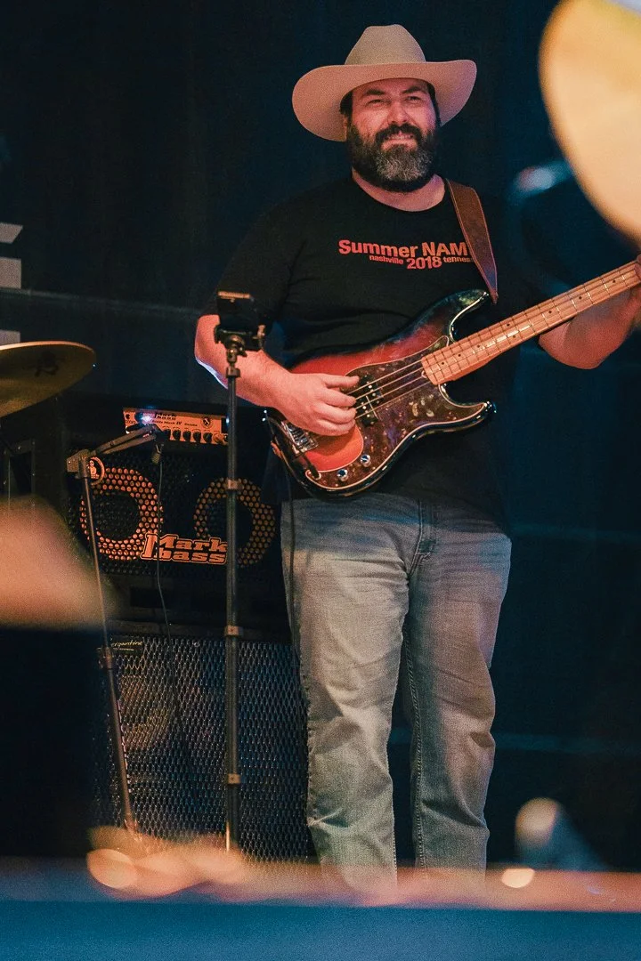 A man with a beard wearing a cowboy hat, black T-shirt, and jeans, playing an electric guitar on stage.