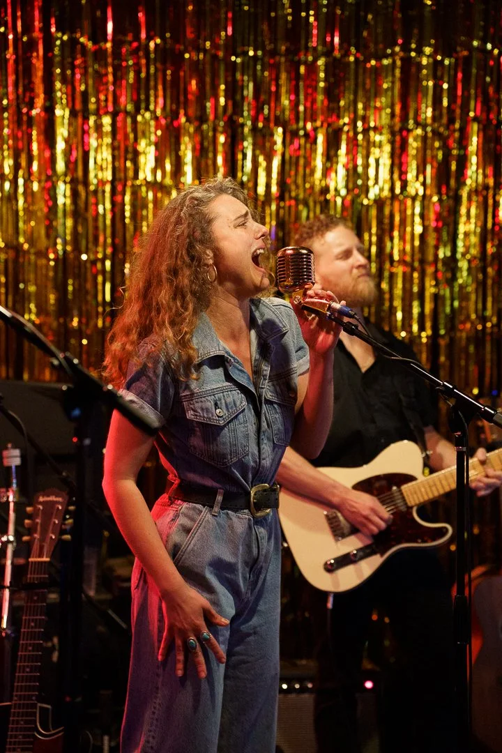 A woman singing passionately into a vintage microphone on stage with a guitarist in the background. The stage has a shiny gold and red curtain backdrop, creating a lively concert atmosphere.