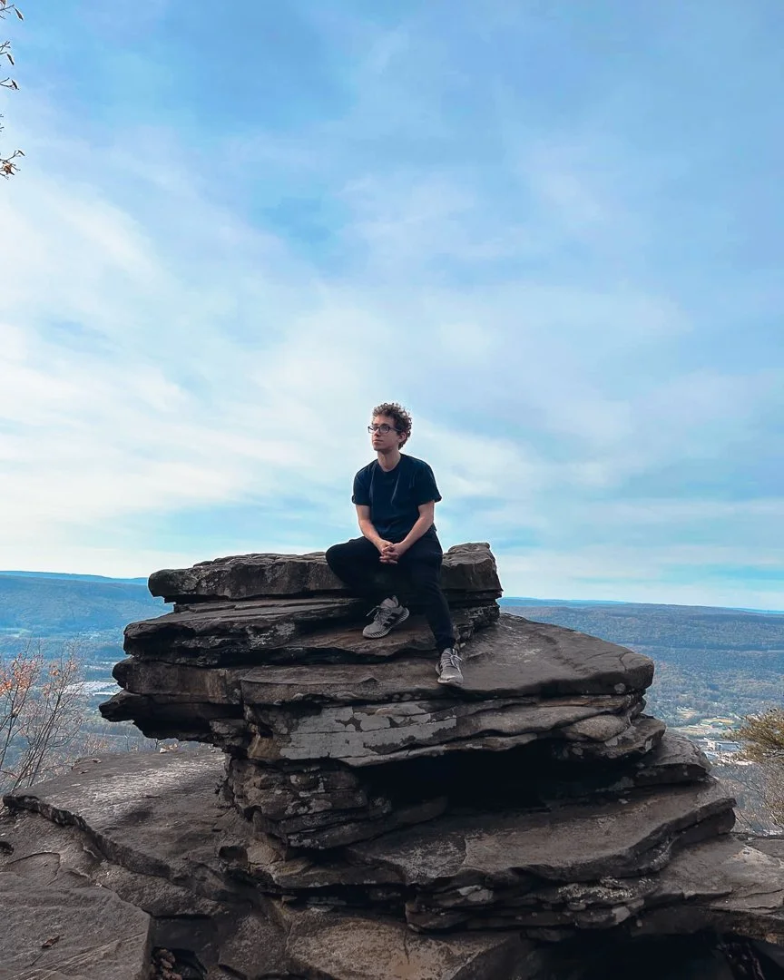 Person sitting on a layered rock formation outdoors, with a scenic view of the landscape and sky in the background.