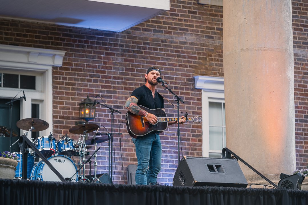 A man playing guitar and singing into a microphone on an outdoor stage with a brick building in the background.