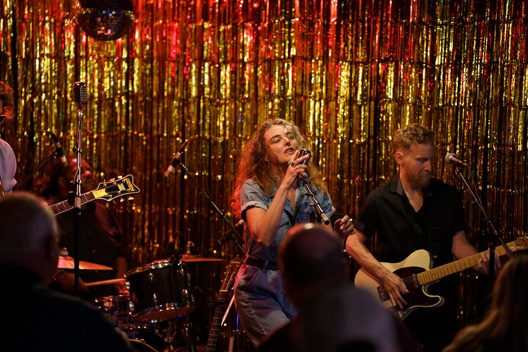 A band performing on a stage decorated with gold and red metallic streamers. A female singer is at the center, holding a microphone, with a guitarist and drummer surrounding her. Audience members are visible in the foreground.