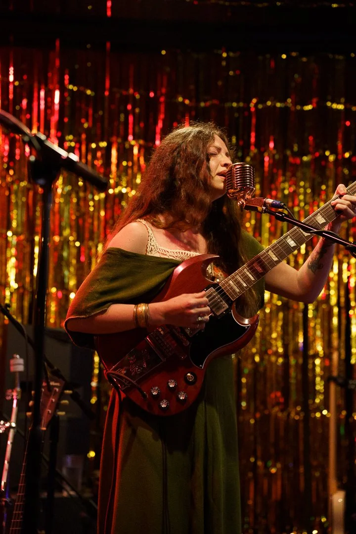 A woman singing into a microphone and playing an electric guitar on stage with a shiny, gold, tinsel backdrop.
