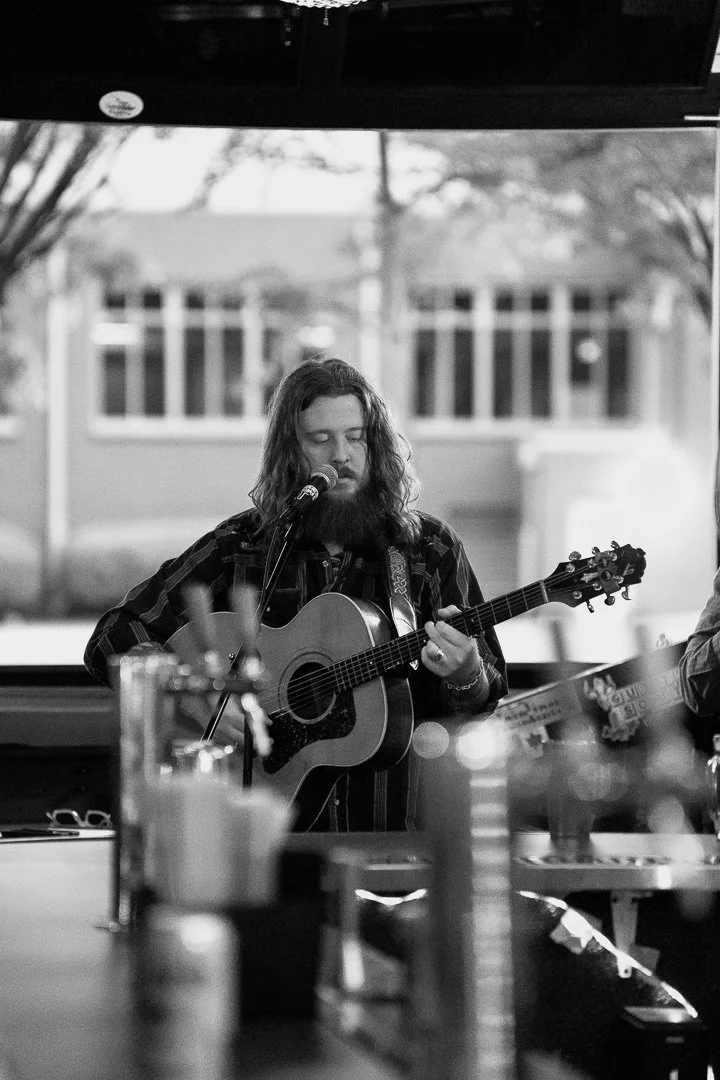 A man with long hair and a beard playing an acoustic guitar and singing into a microphone in a room with large windows.
