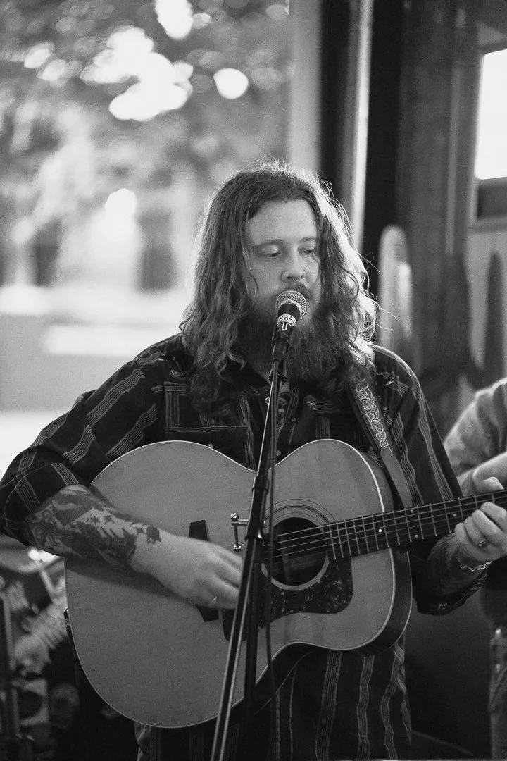 A woman with long wavy hair and tattoos on her arm is singing into a microphone while playing an acoustic guitar in a cozy indoor setting.