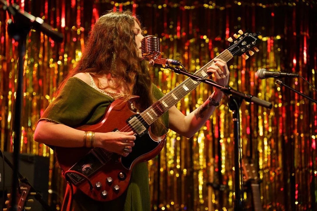 A female musician performing on stage with an electric guitar, singing into a vintage microphone, in front of a gold and red tinsel backdrop.