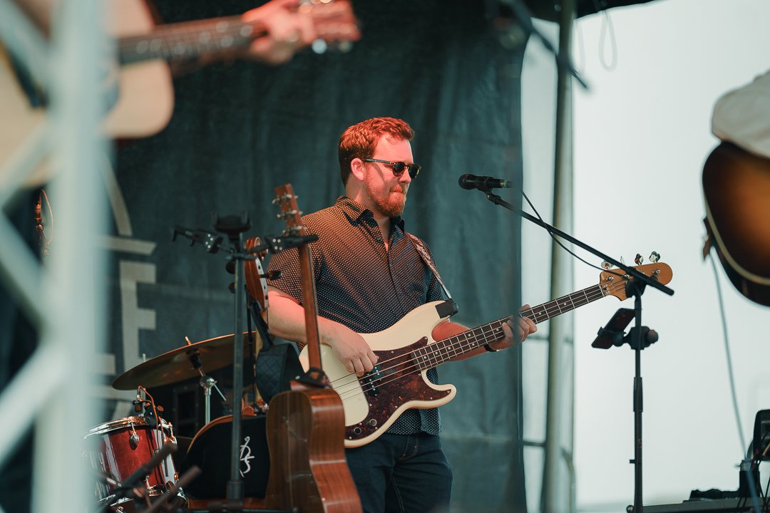 A man with sunglasses and a beard playing a bass guitar on stage at a music festival.