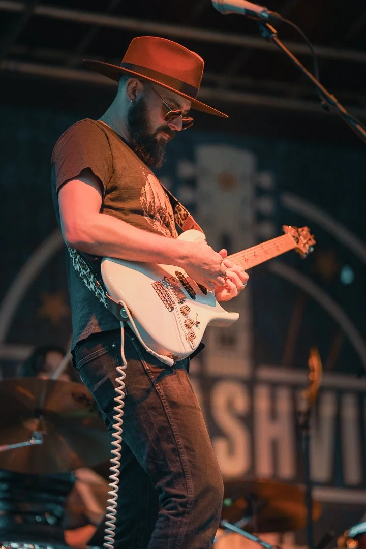 A man wearing a wide-brimmed hat, sunglasses, and casual clothing playing an electric guitar on stage.
