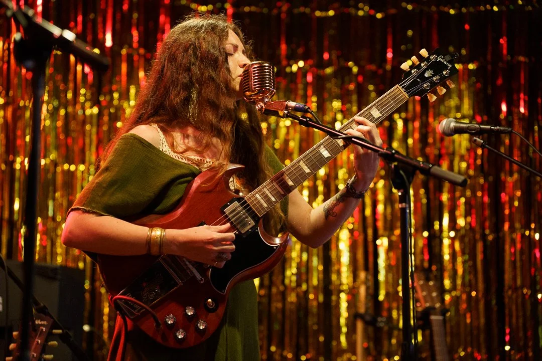 A woman with long curly hair playing an electric guitar and singing into a retro microphone on stage, with a gold tinsel curtain in the background.