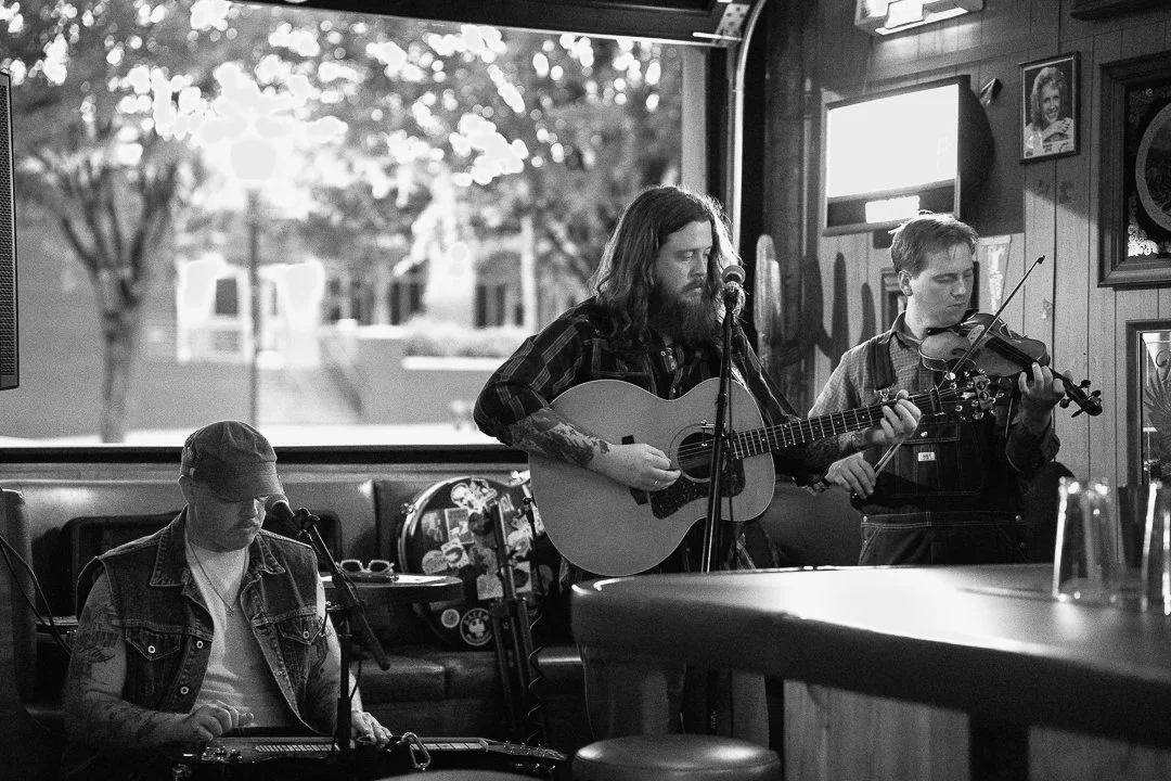 Three musicians performing inside a bar with a window showing trees outside; one playing the keyboard, one with a guitar, and one on the violin.