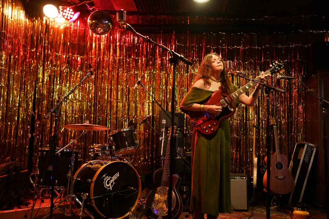 A woman performing on stage with a guitar, surrounded by musical instruments, against a shimmering metallic backdrop.