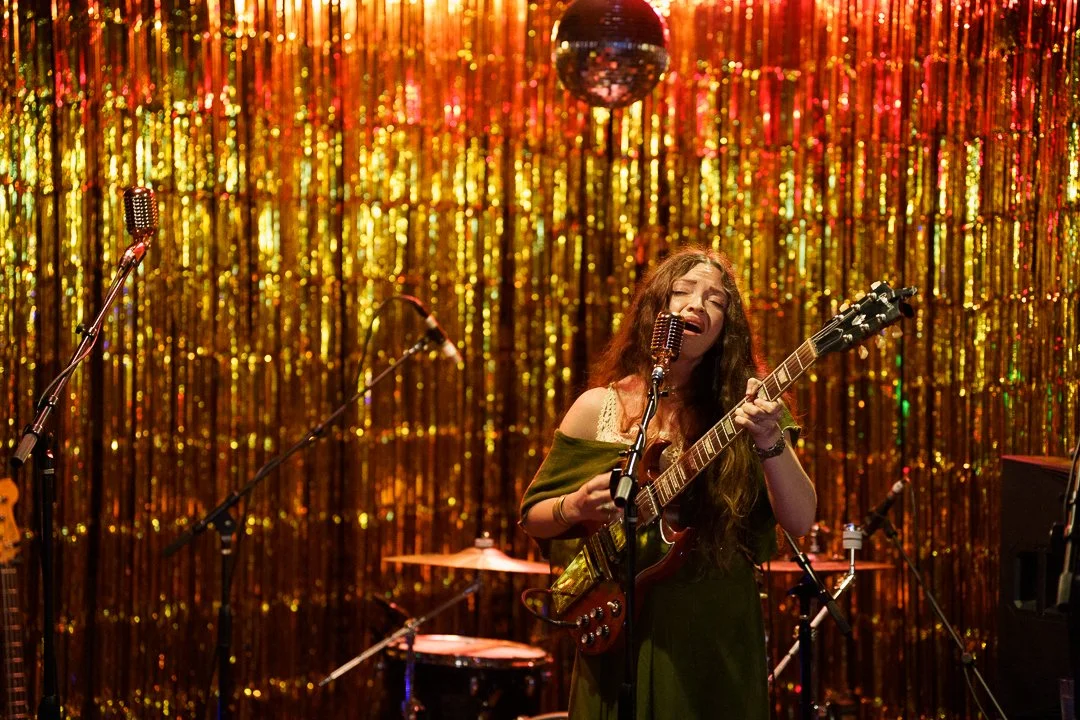 A young woman with long hair singing and playing guitar on a stage with a gold shimmering curtain background and disco ball overhead.