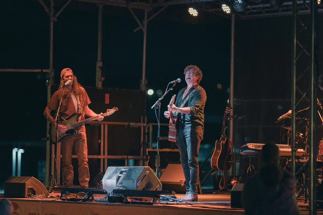Two musicians performing on stage at night, one playing guitar and singing into a microphone, and the other with a guitar and wearing a hat.