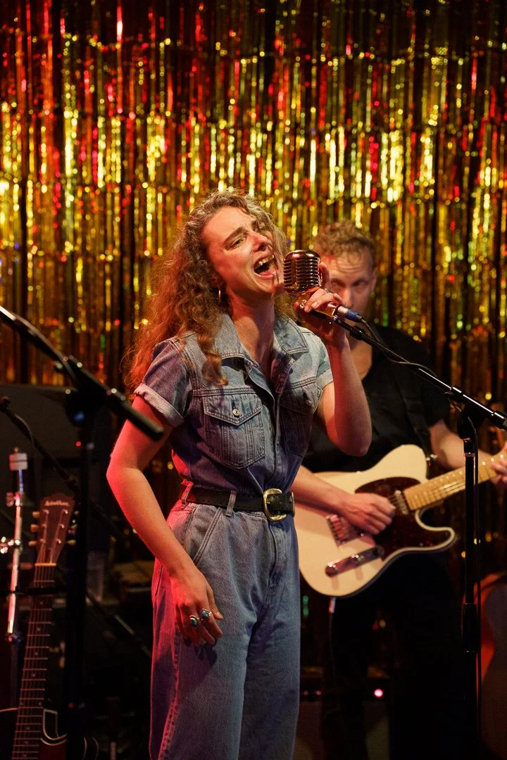 Woman singing passionately into a vintage microphone on stage with gold tinsel backdrop, accompanied by a guitarist.