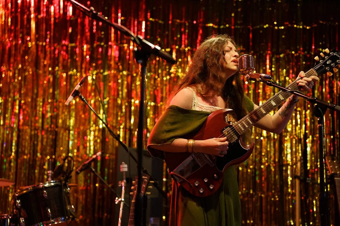 A woman with long curly hair playing an electric guitar and singing into a vintage microphone on a stage with red and gold tinsel backdrop.