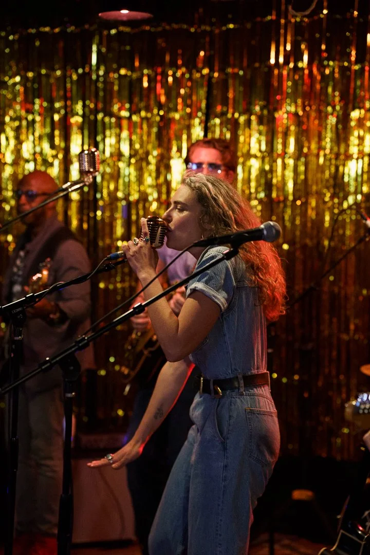 A woman singing into a vintage microphone on stage; she has curly hair, wearing a denim outfit, and there are musicians and a shiny gold background behind her.