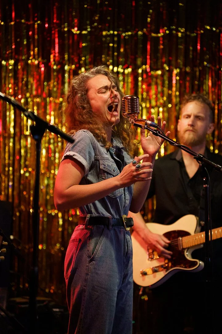 A woman singing passionately into a vintage microphone on stage, with a guitarist in the background, in front of a shiny, gold, tinsel curtain.