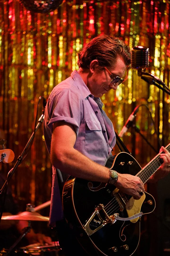Man playing an electric guitar on stage with a shiny, gold and red tinsel curtain backdrop.