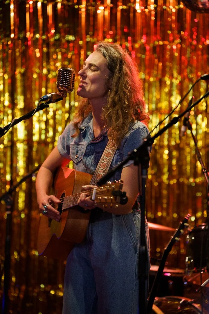 A woman with wavy hair wearing a denim shirt and jeans, singing into a vintage microphone while playing an acoustic guitar on stage with a gold sequin backdrop.