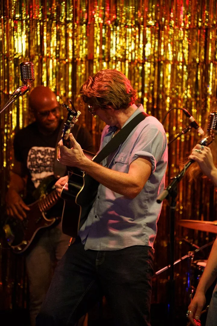 Musicians performing on stage, with a man playing an acoustic guitar, and another man in the background playing an electric guitar, against a gold glitter curtain backdrop.