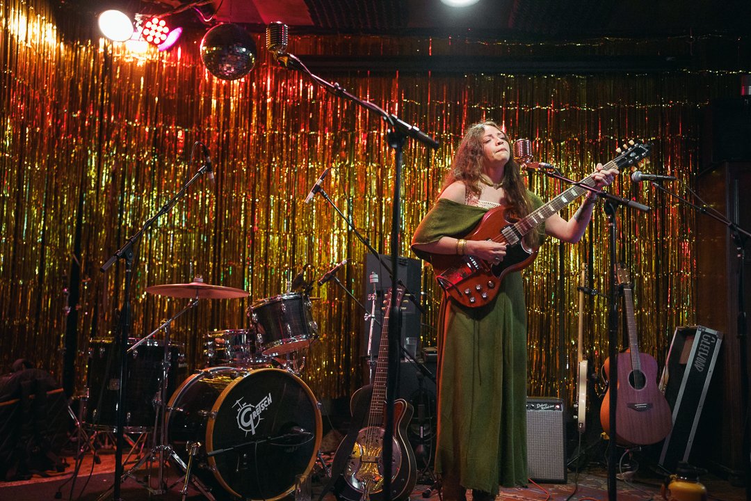 A woman with long hair singing and playing guitar on stage, decorated with gold tinsel backdrop, with drums and other guitars around.