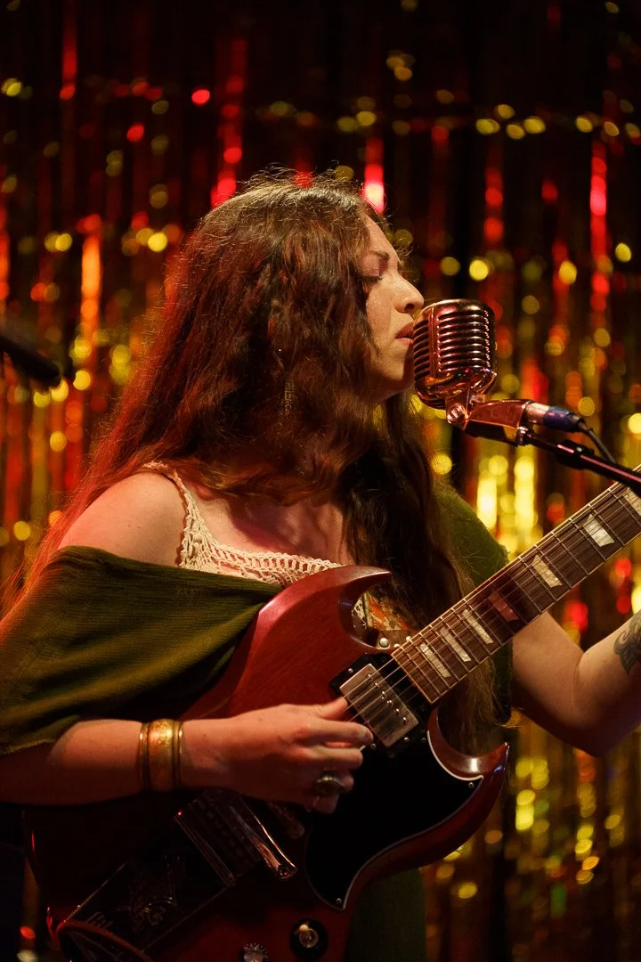 A woman with long, curly hair singing into a vintage microphone while playing an electric guitar during a performance with a sparkling gold and red curtain in the background.
