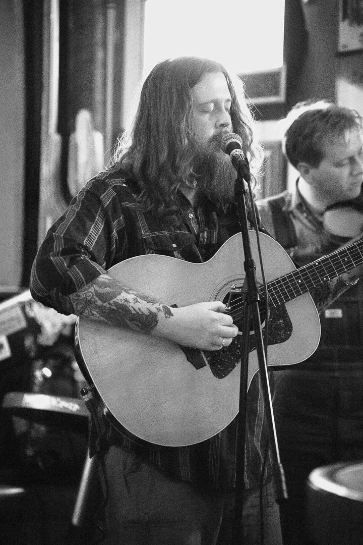 A man with long hair, a beard, and tattoos on his forearm is singing into a microphone while playing an acoustic guitar in a dimly lit room, with another person playing the drums in the background.