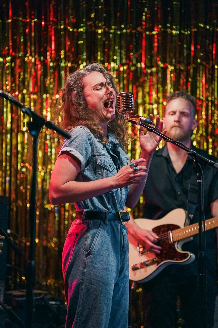 A woman with curly hair singing passionately into a vintage microphone on stage with a man playing an electric guitar in the background, against a shimmering gold backdrop.