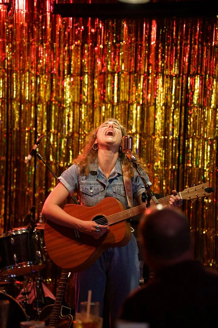 A woman singing and playing guitar on stage with a gold and red tinsel backdrop.