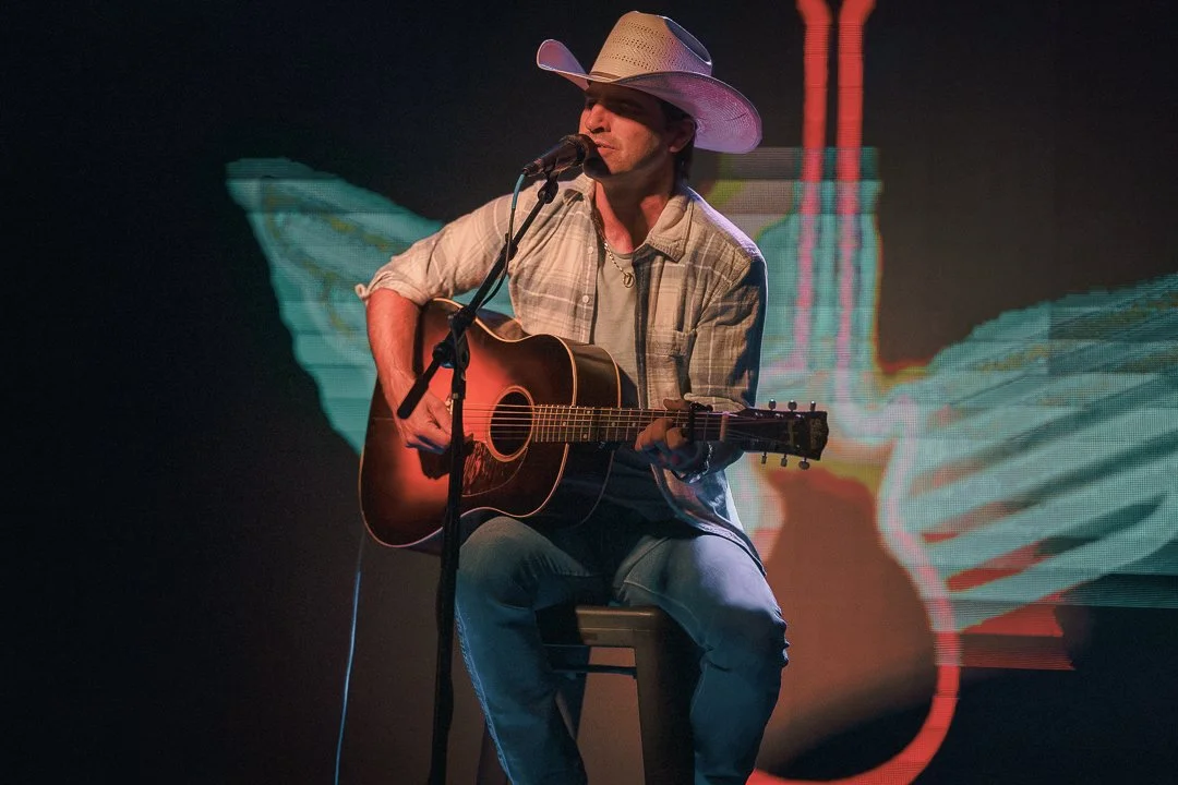 A man wearing a cowboy hat is playing an acoustic guitar and singing into a microphone on stage.