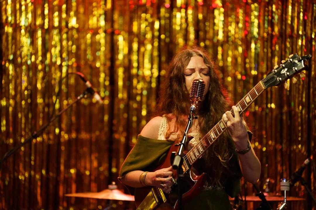 A woman with long wavy hair sings into a vintage microphone while playing an electric guitar on stage, with a glittery gold backdrop.