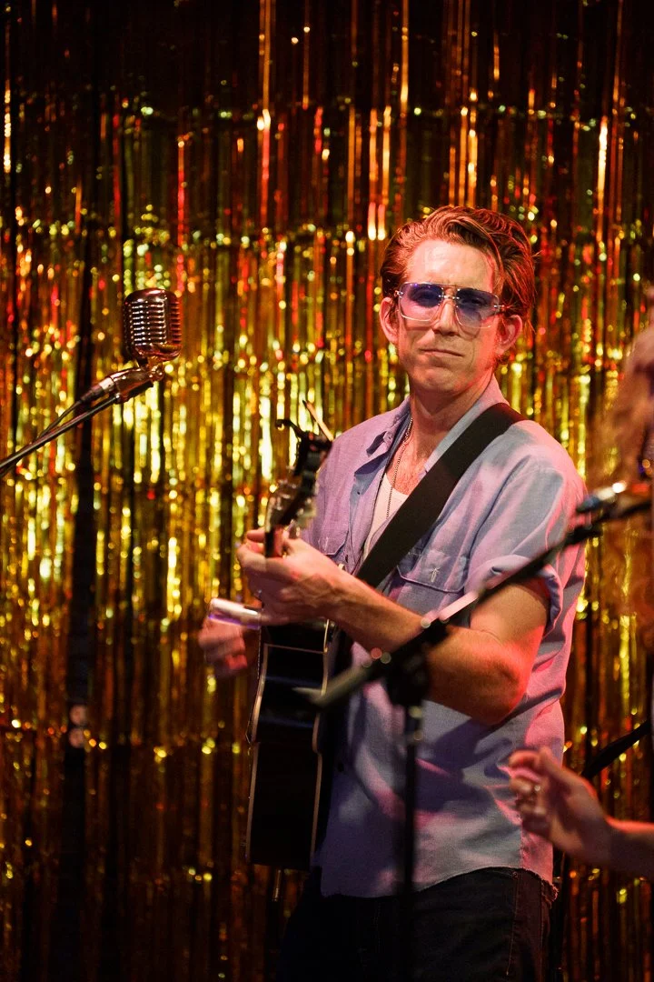 A man with red hair and sunglasses playing an acoustic guitar on stage with a gold tinsel backdrop.