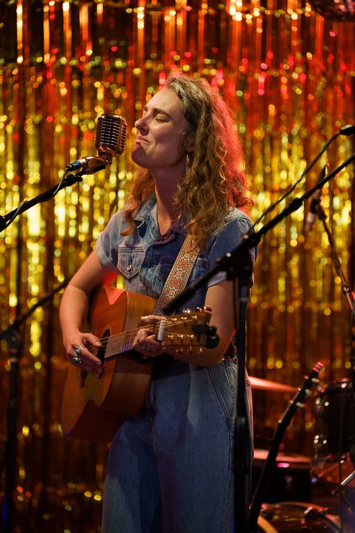 A woman with curly hair playing an acoustic guitar on stage, singing into a vintage microphone with a golden, glittery curtain in the background.