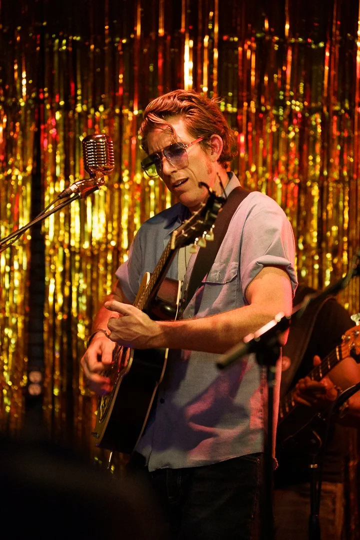 Man with wavy hair wearing sunglasses playing acoustic guitar on stage with gold tinsel backdrop.