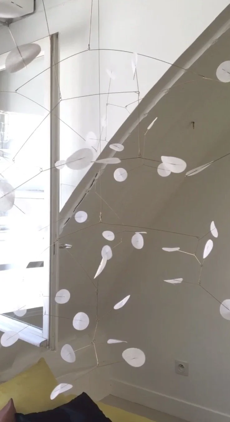Installation artistique suspendue avec des cercles blancs en papier ou en plastique, accrochés à des fils fins, dans une pièce avec une fenêtre claire et un mur blanc.