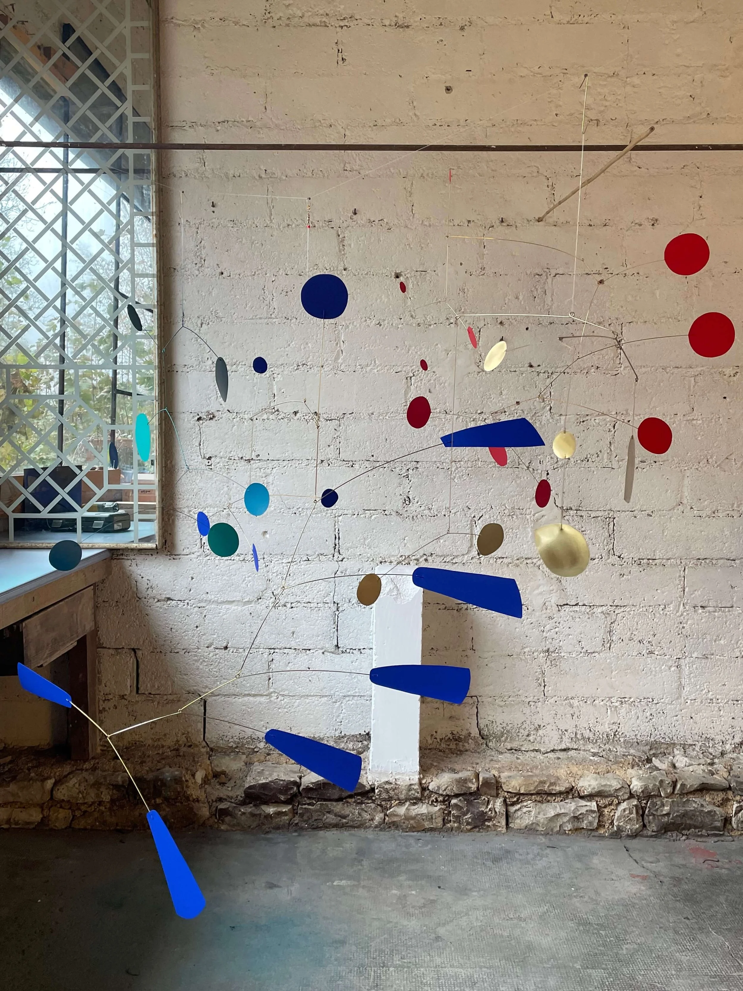 Mobile d'art abstrait avec des formes ovales et rectangulaires colorées en bleu, rouge, or, blanc, gris, turquoise, suspendu dans un espace avec un mur en pierre et une fenêtre avec grille