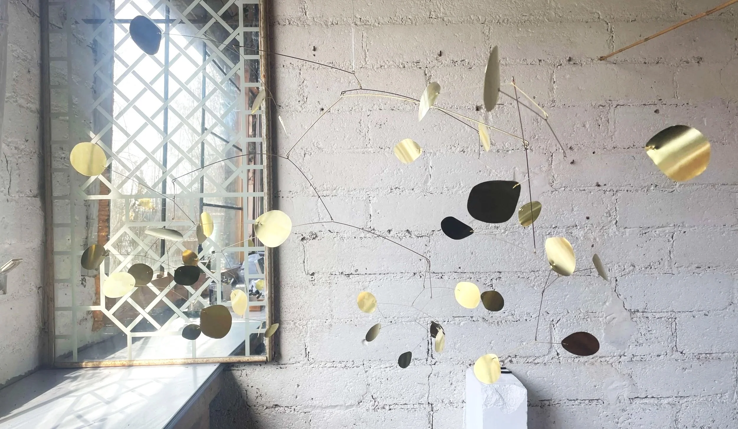 Une sculpture suspendue de disques métalliques dorés et noirs dans une pièce avec un mur blanc en briques et une fenêtre.