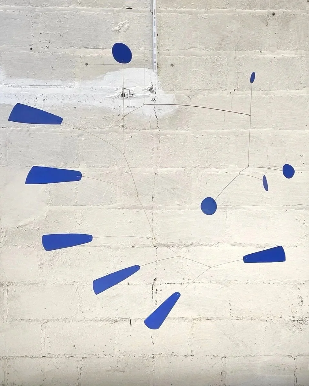 Une œuvre d'art abstraite utilisant des formes bleues en papier ou en plastique fixées à un mur en brique blanc, assemblées de manière à évoquer l'horloge ou la constellation.
