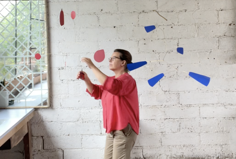 Une femme portant un chemisier rouge et des lunettes joue avec des éléments décoratifs suspendus devant un mur en briques blanches, à côté d'une fenêtre avec un quadrillage en bois.
