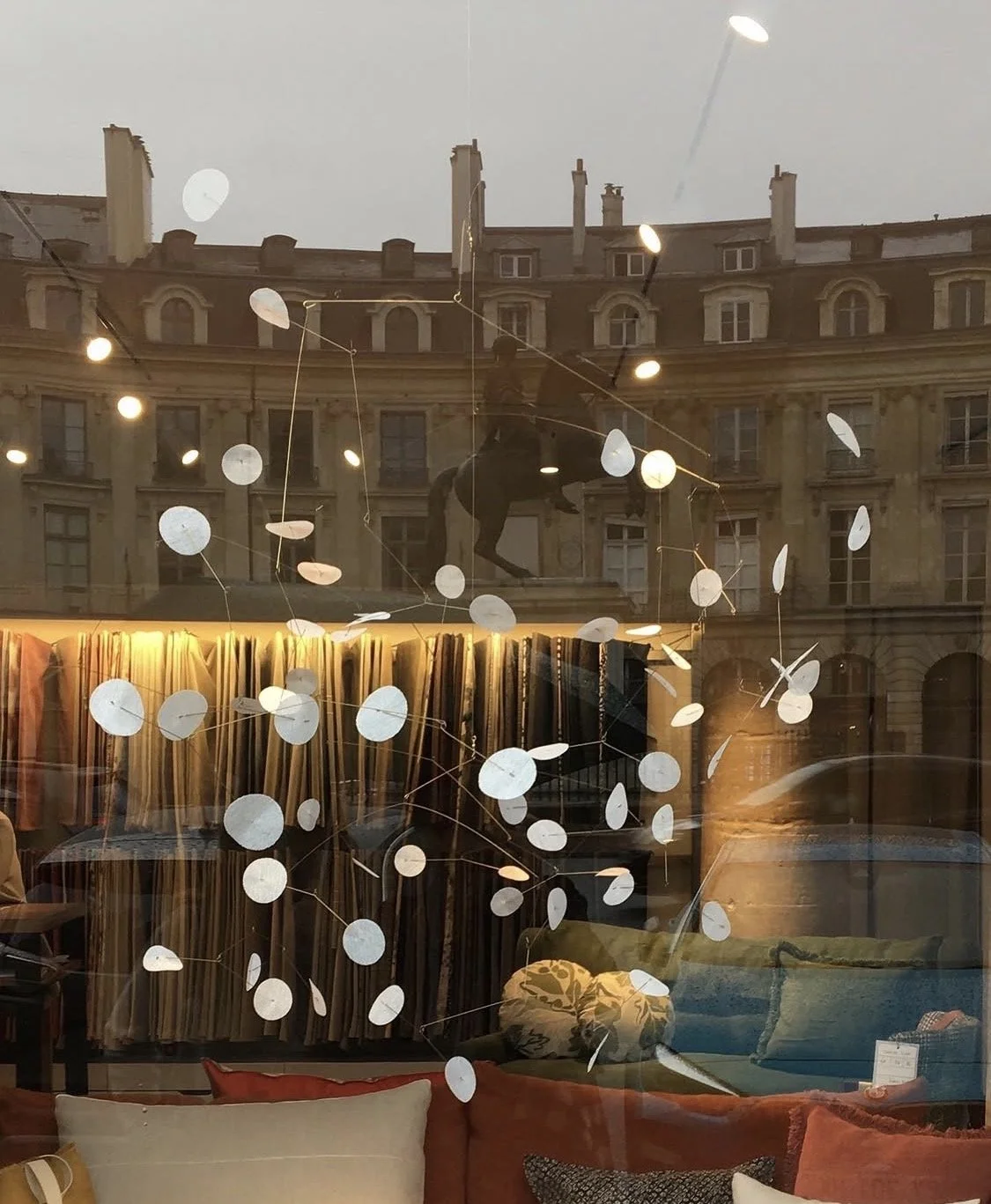 Reflet d'un bâtiment avec fenêtres et un cheval en silhouette, ainsi qu'une décoration suspendue en forme de disques blancs dans une vitrine d'ameublement.