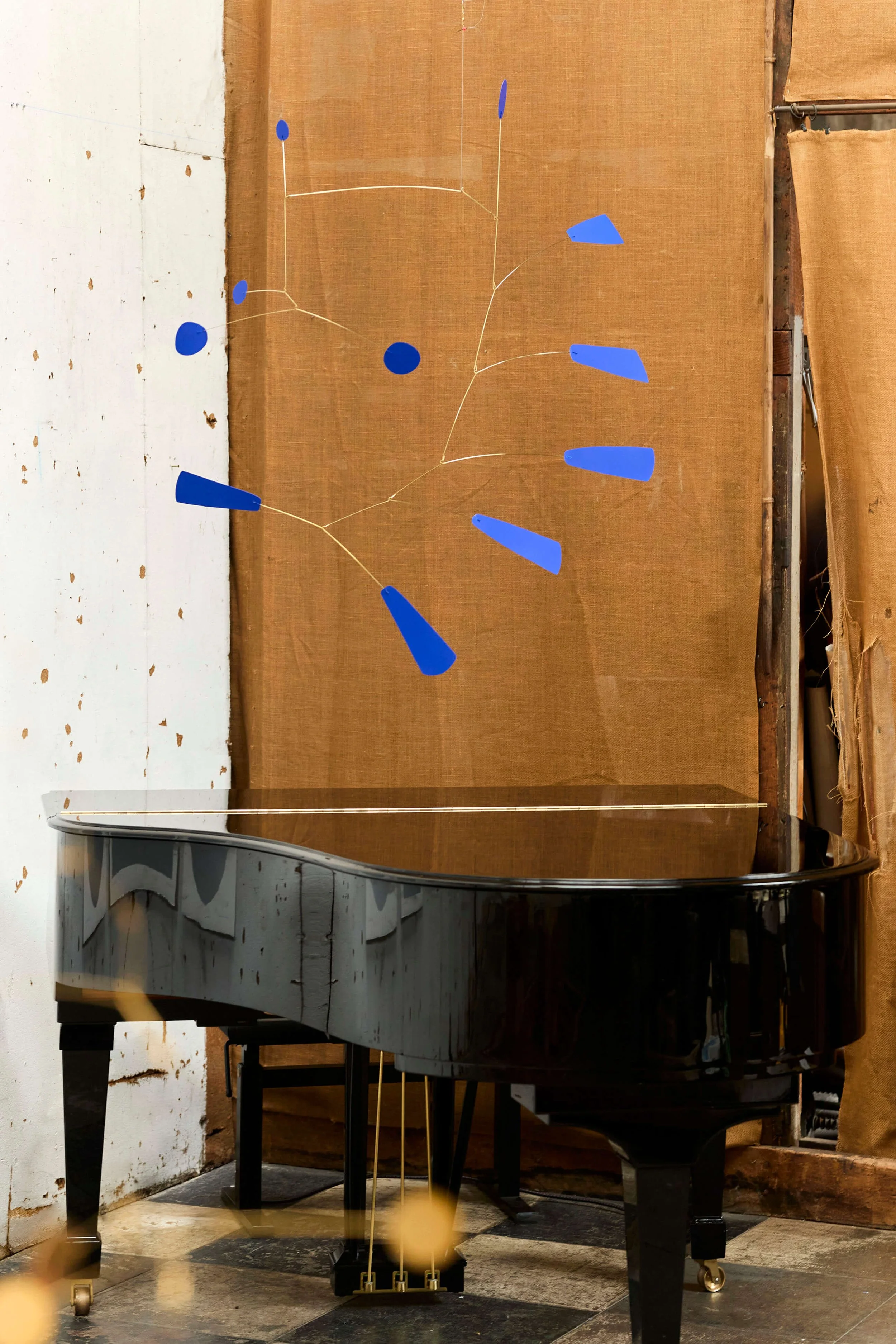 Un piano à queue noir dans une pièce avec des murs en bois et un sol en carreaux. La photo est prise de côté, montrant ses pédales en métal et ses roulettes en dessous.