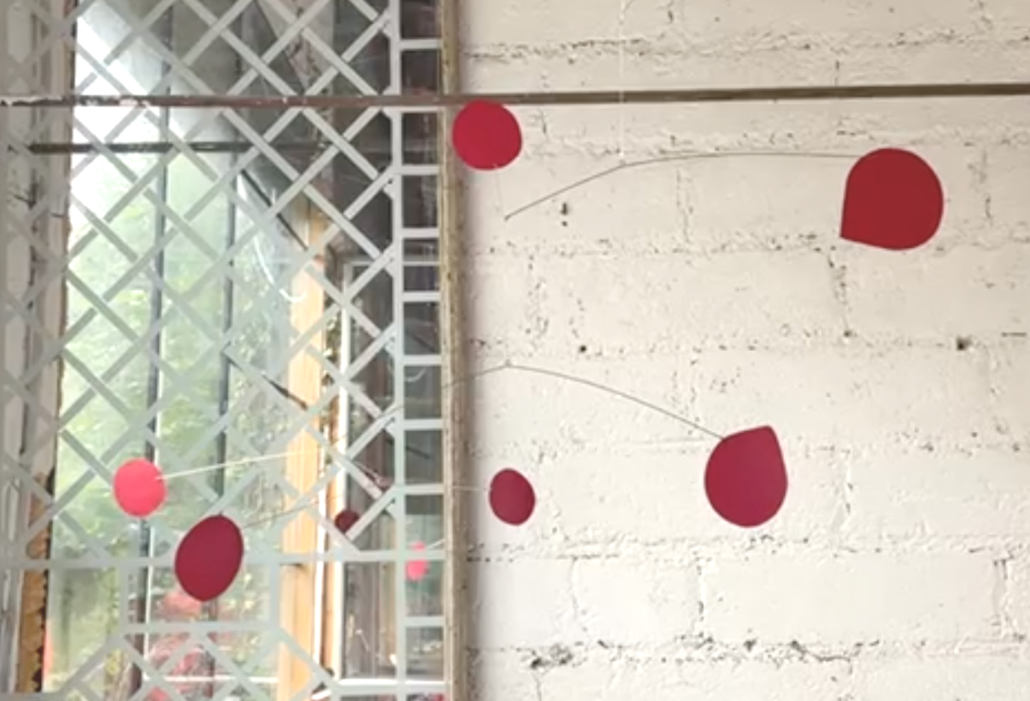 Installation artistique avec des sphères rouges suspendues par des fils fins devant une grille blanche et un mur blanc.