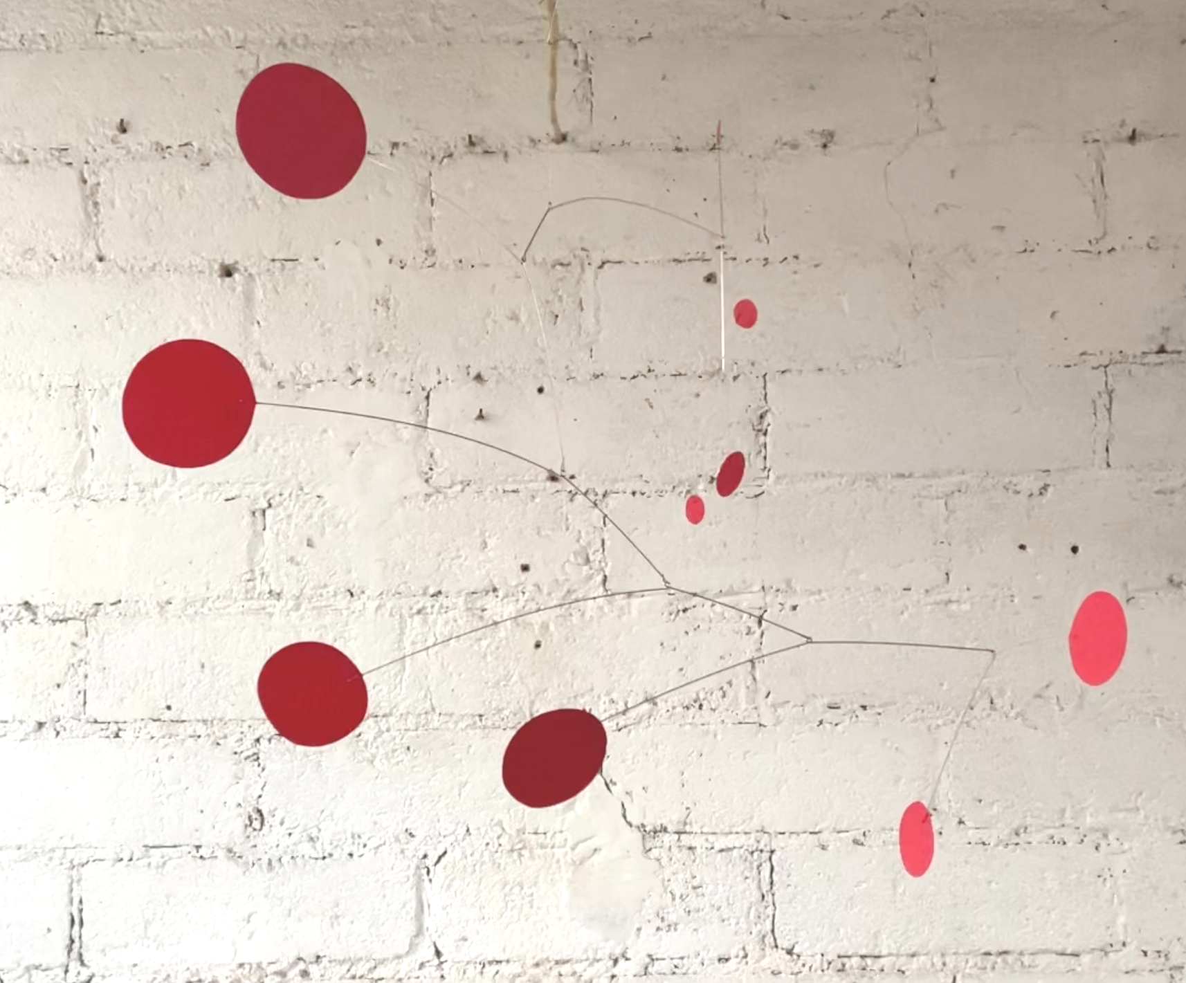 Ceci est une œuvre d'art abstraite représentant plusieurs cercles rouges de différentes tailles, reliés par de fines lignes, placé sur un mur de briques blanches.