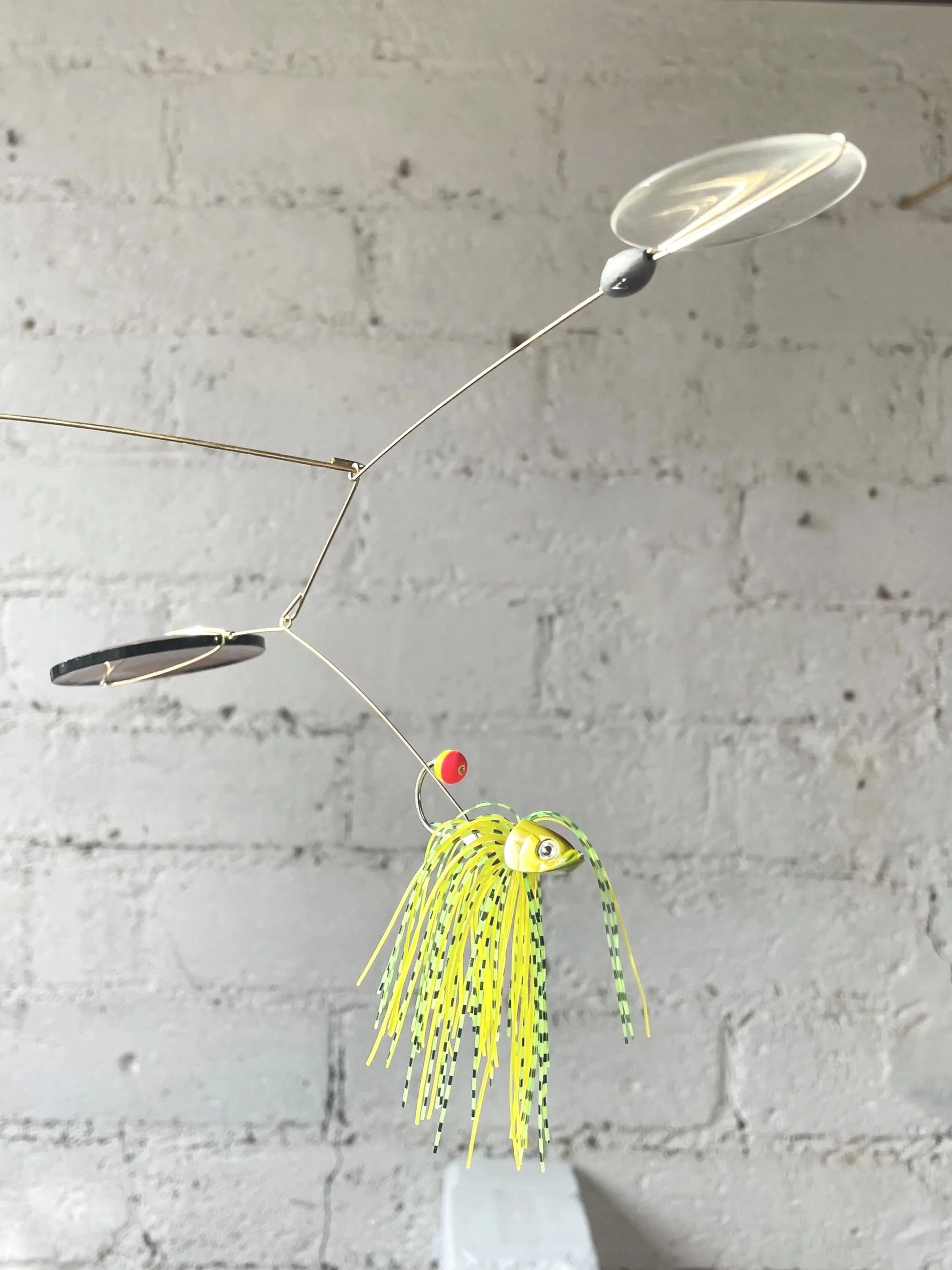 Une décoration suspendue en forme de poisson jaune avec des nageoires en tresses, suspendue par un fil à une structure métallique. La structure comprend des éléments en plastique, une boucle avec une bille noire, une feuille en plastique transparent, et une petite boule rouge. Le fond est un mur en briques grises.