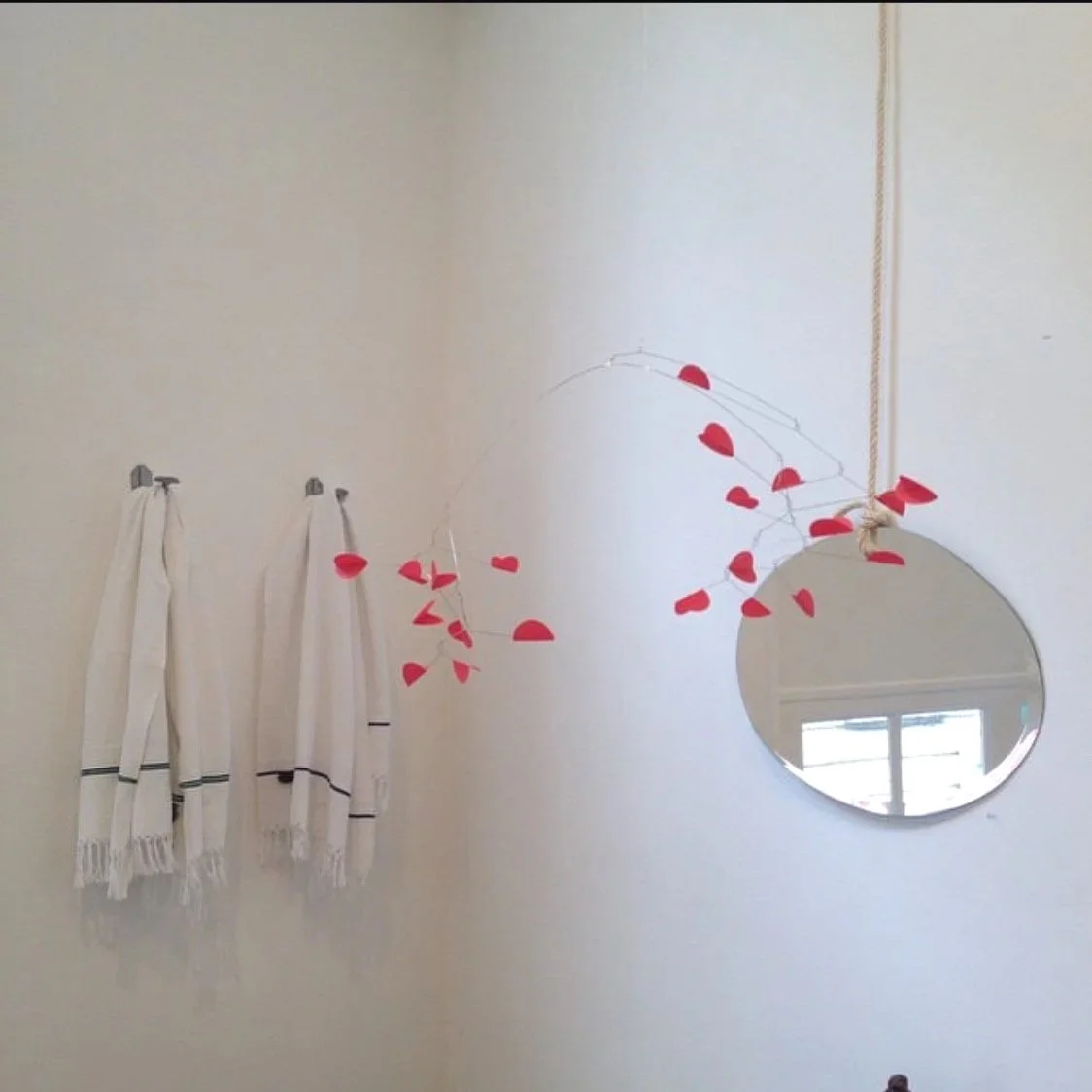 Décoration murale avec un miroir rond suspendu par une corde, entouré de guirlandes de coeurs rouges et blanche, avec des serviettes blanc à rayures noires accrochées au mur.