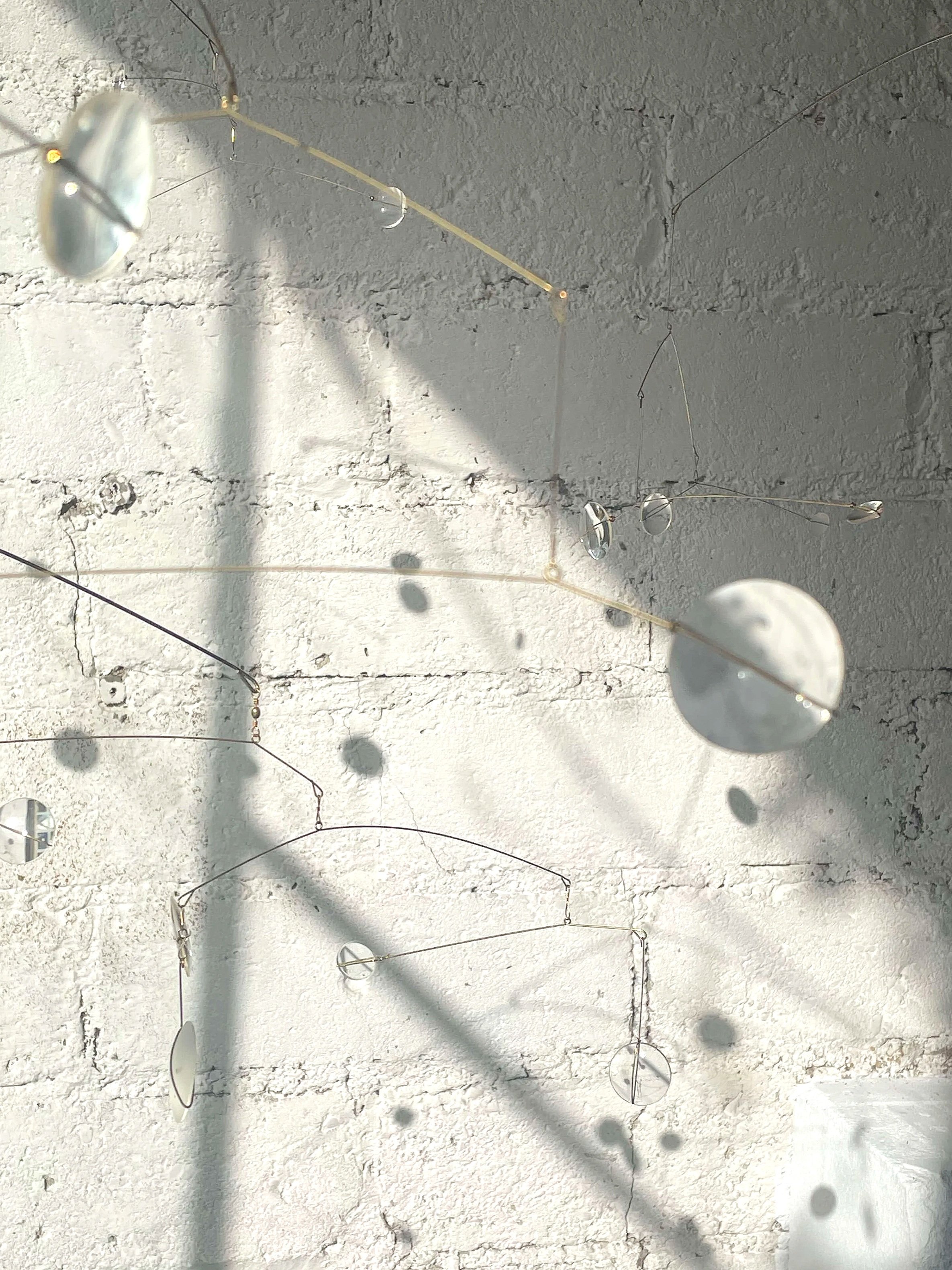 Une installation artistique suspendue composée de plusieurs miroirs ronds fixés à des fils fins, projetant des ombres sur un mur en pierre blanc.