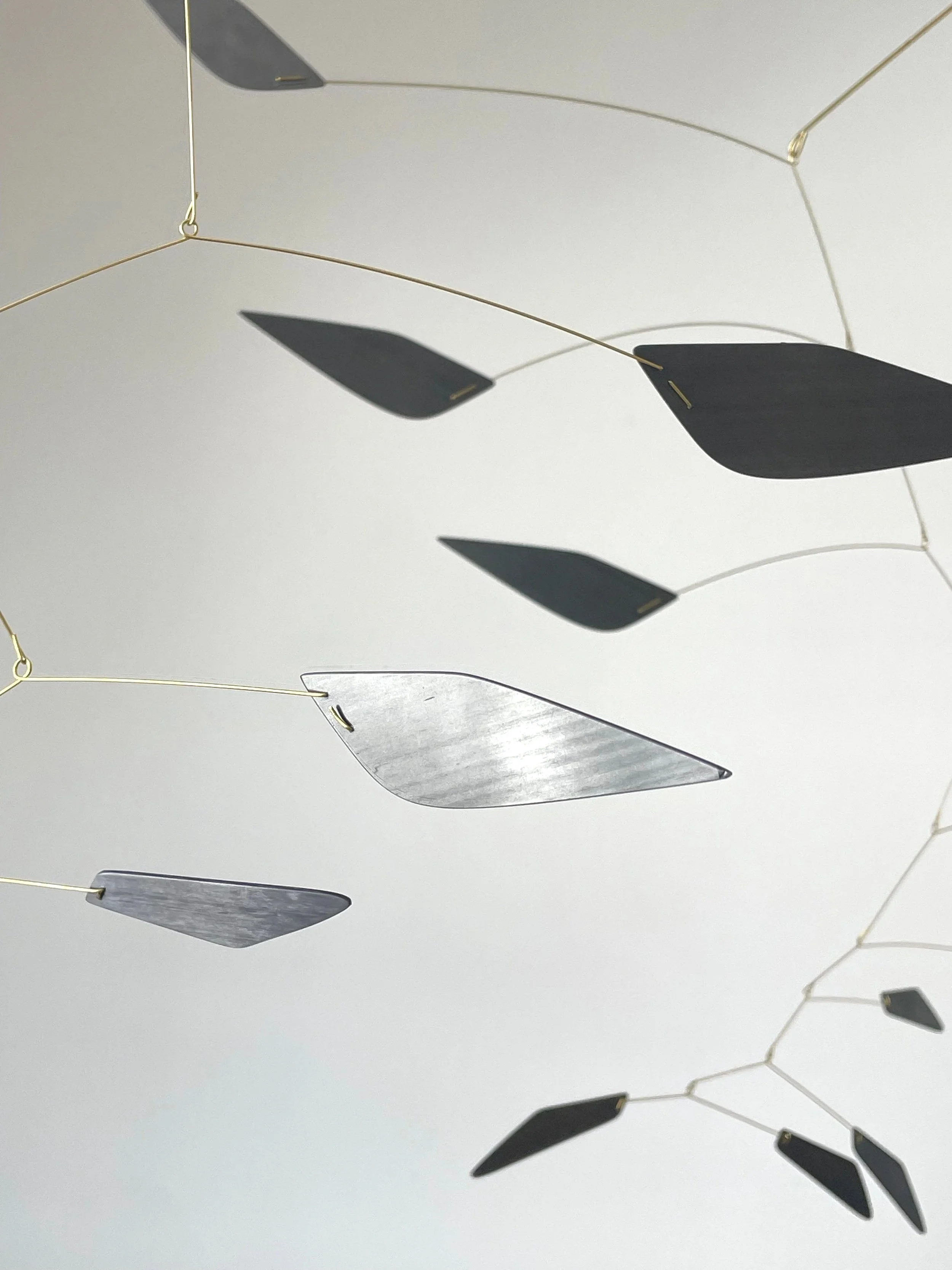 Une décoration suspendue faite de plusieurs éléments en forme de feuilles en métal noir et gris, attachés à des fils fins, suspendus contre un plafond blanc.