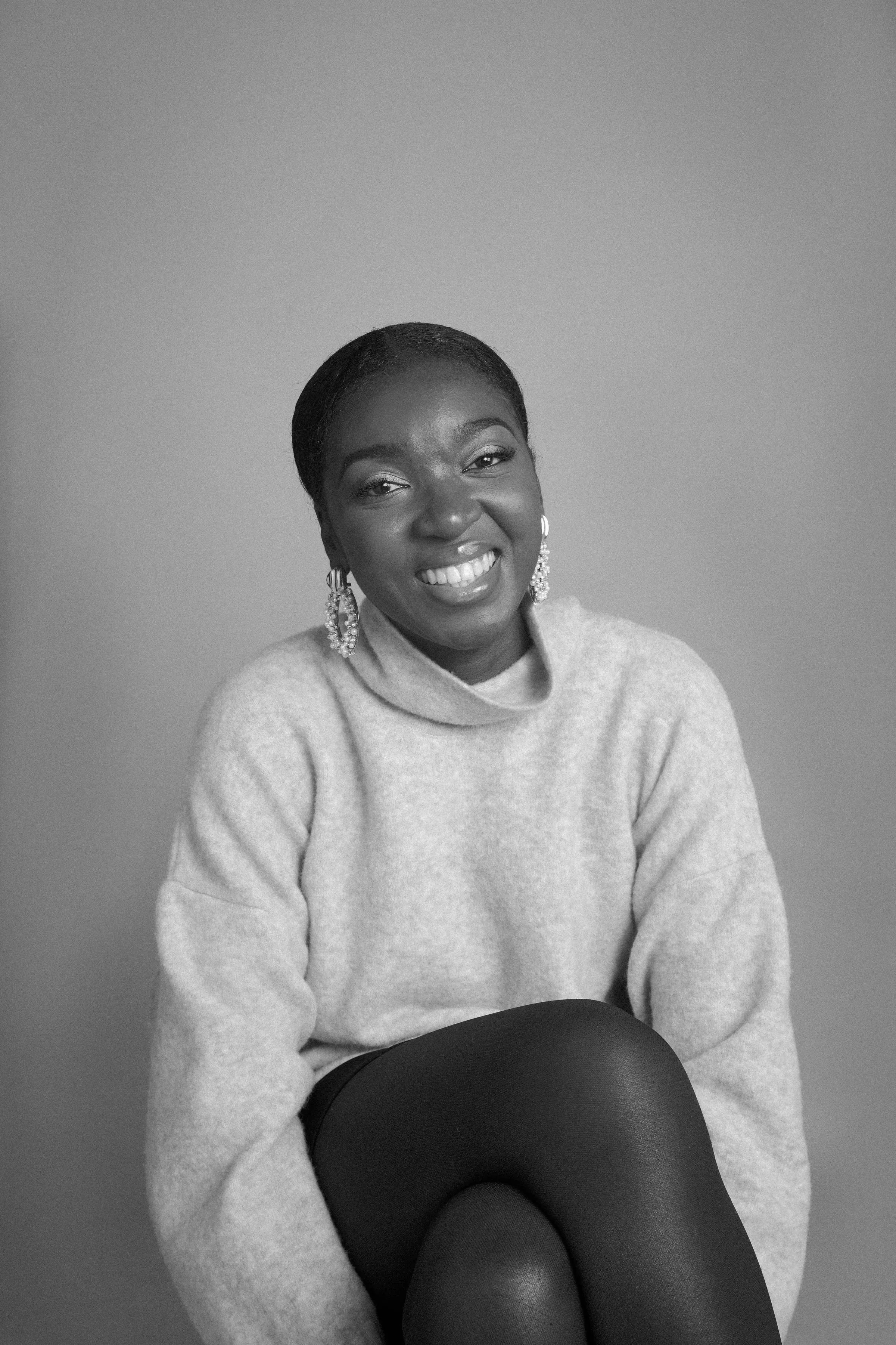Black and white portrait of a woman smiling, wearing earrings and a light sweater, sitting with legs crossed against a plain background.