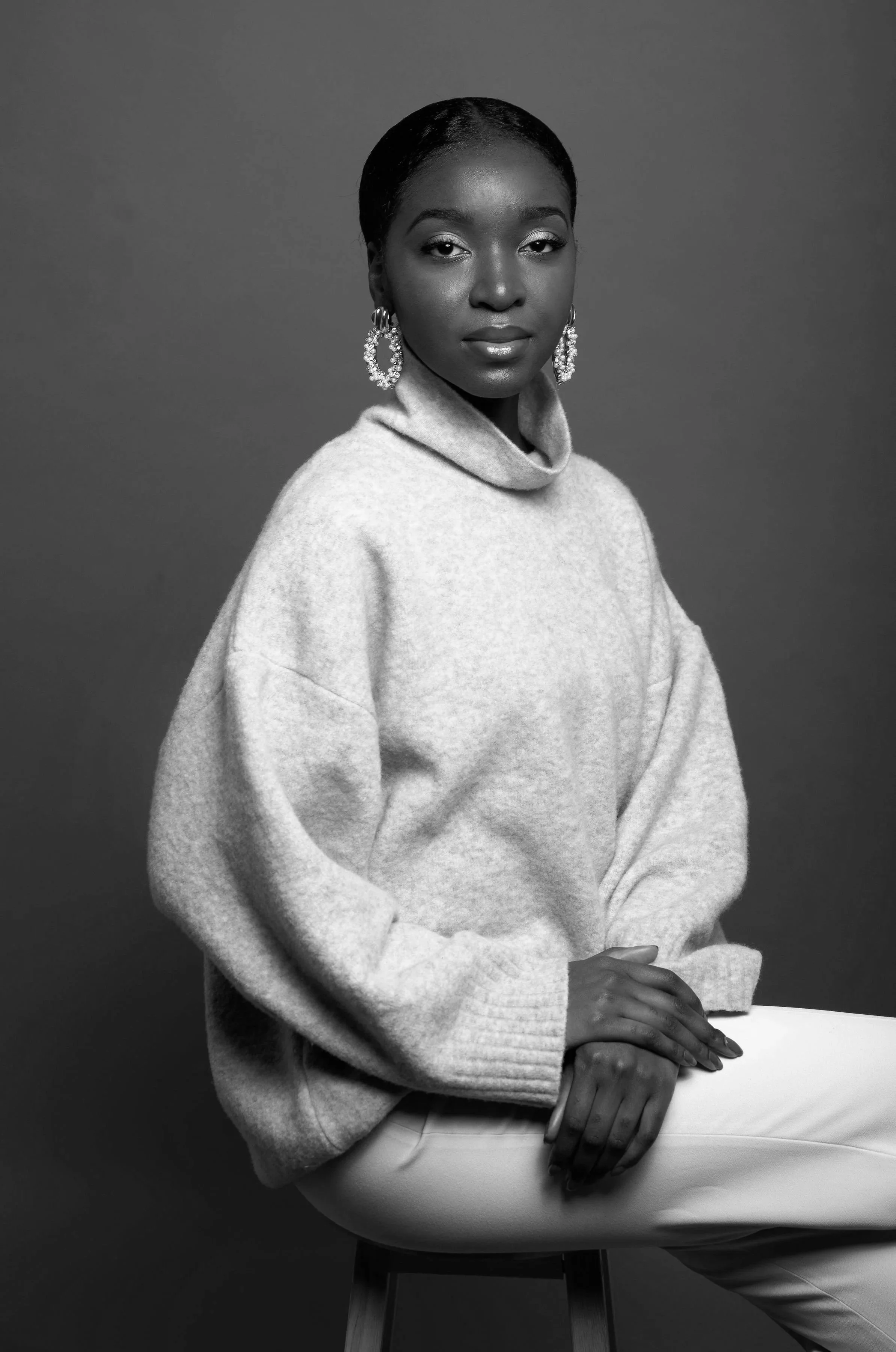 Black and white portrait of a woman with short hair, wearing statement earrings and a high-neck sweater, seated and resting her hands on a table. She is looking directly at the camera with a calm expression.